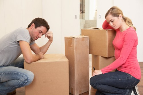 A young man and a young woman are indoors, surrounded by several cardboard boxes of varying sizes, likely in a residential setting preparing for a house move in the Belgravia area. The man, dressed in a grey t-shirt and jeans, is sitting on the floor with one elbow resting on a box, appearing contemplative or concerned, while the woman, wearing a bright pink long-sleeved top and dark jeans, is kneeling beside a large box, resting her head on her hand and looking down with a thoughtful expression. The boxes are made of plain brown cardboard, some sealed with packing tape, and show signs of recent packing with crumpled flaps and labels. The background reveals a light-colored wall and a staircase, indicating the interior of a house. The natural lighting highlights the textures of the cardboard, fabric clothing, and wooden flooring. This scene aligns with house removal activities typical of local moving services in Belgravia, emphasizing the careful handling and packing of household belongings associated with professional removals.