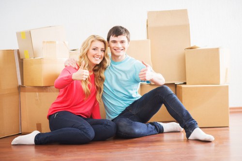 A young couple sitting on a wooden floor in a residential property with light-colored walls, surrounded by stacked cardboard moving boxes of varying sizes, some sealed with packing tape. The woman, with long blonde hair, is wearing a red long-sleeve top and dark jeans, smiling and giving a thumbs-up with her right hand. The man, with short dark hair, is dressed in a light blue T-shirt and dark jeans, also smiling and giving a thumbs-up with his left hand. They appear to be in a hallway or living room, with natural light illuminating the scene, indicating a house or flat in the UK, possibly within the Belgravia or nearby postcode areas. The scene reflects a positive moment during a house move, aligned with professional removals services provided by Belgravia Man with Van, highlighting the safe handling and transportation of household belongings in a local residential setting.