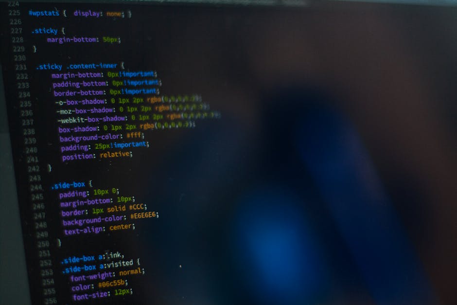 A close-up photograph of a computer screen displaying lines of CSS code in various colors such as green, blue, yellow, and purple, with visible syntax like class selectors and property declarations. T