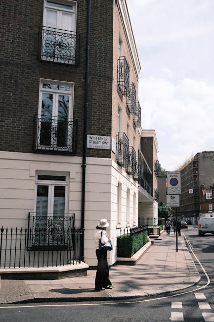 A narrow cobblestone street lined with white terraced houses, each with black or brown doors and windows, some with small balconies or landscaped front gardens. A white van is parked on the right side